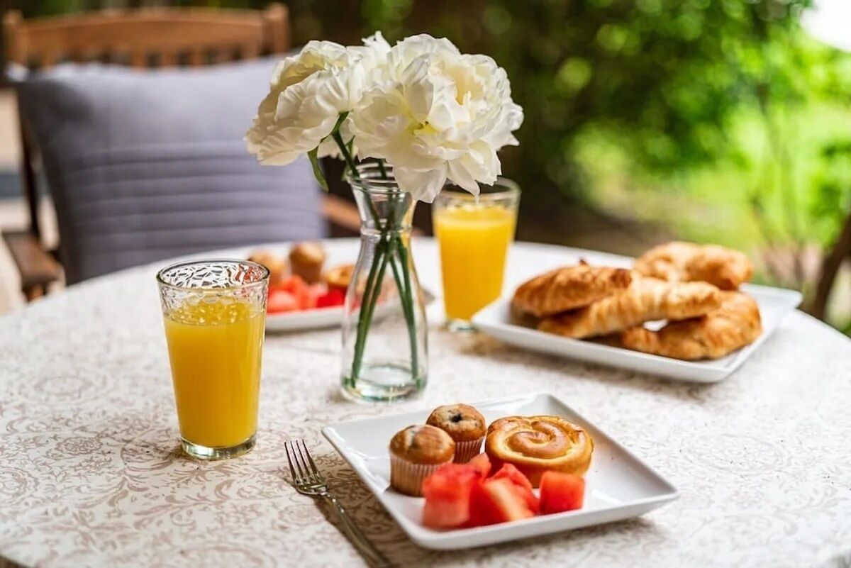 A breakfast setting with croissants, muffins, watermelon, and orange juice on a table with a vase of flowers.