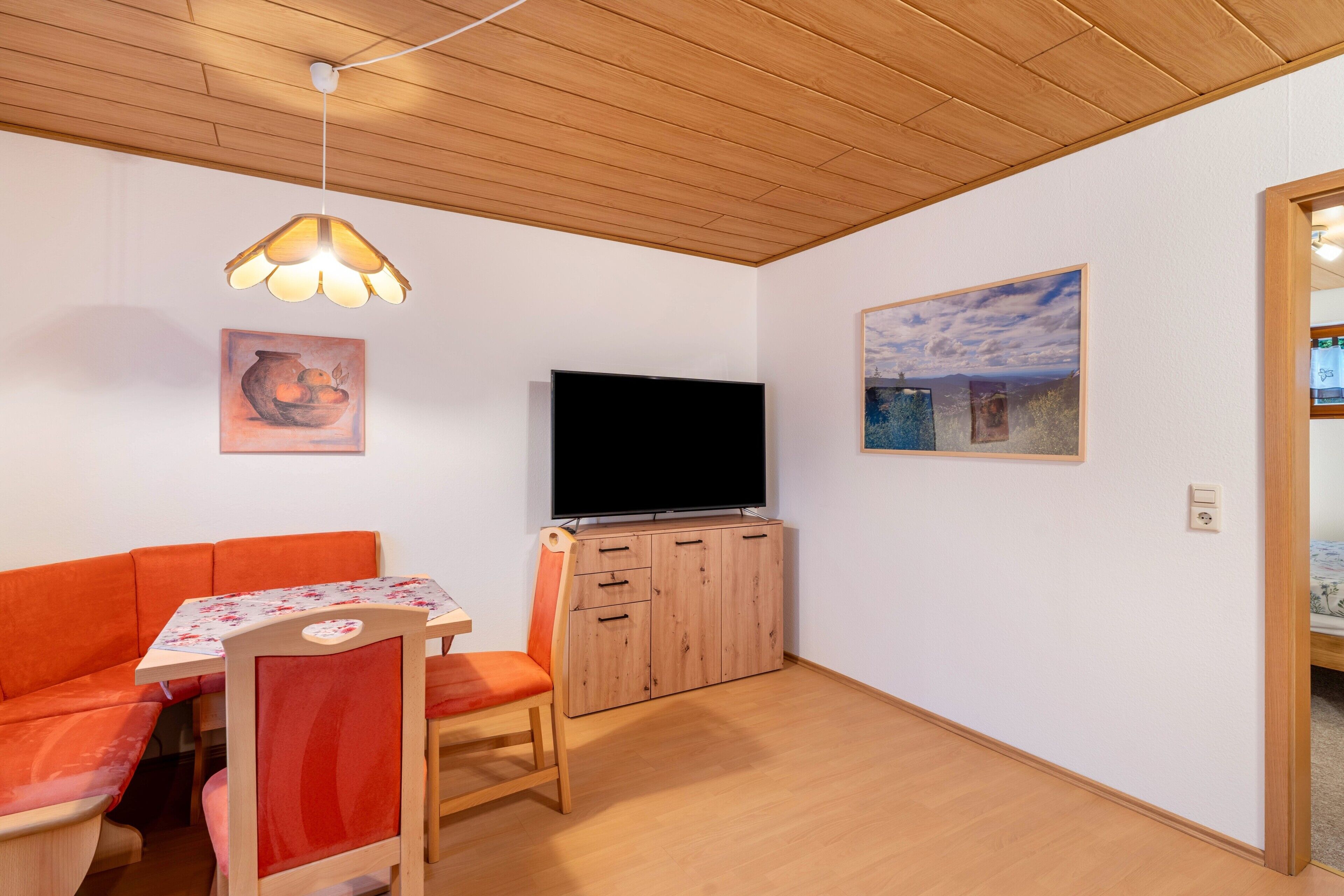 A room with a wooden ceiling, a flat-screen TV, a sofa, a table with a floral tablecloth, and a cabinet.