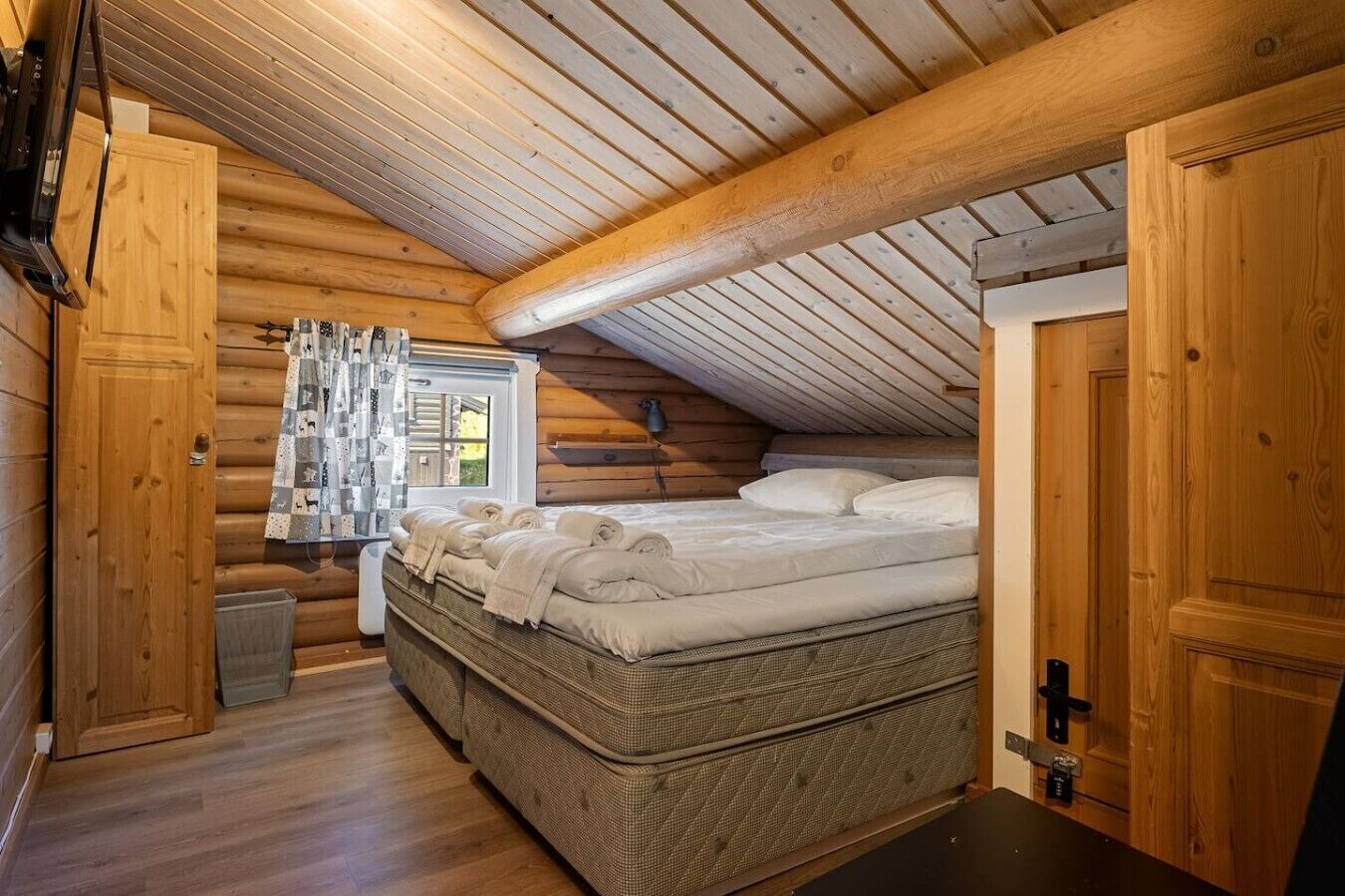 A log cabin bedroom with a bed, wooden walls, a window with curtains, and a door.