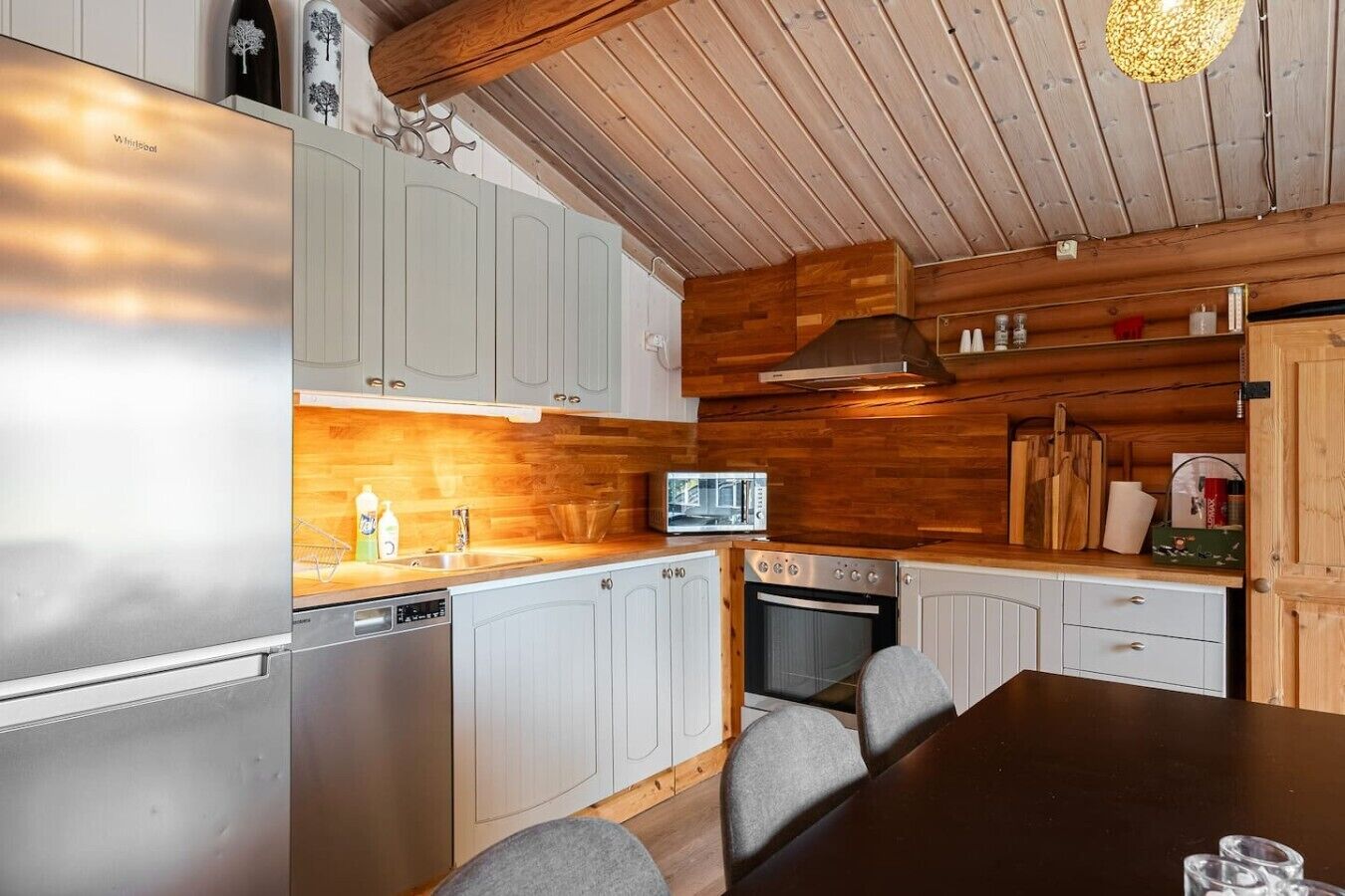 A modern kitchen with a stainless steel refrigerator, a microwave, and a built-in oven. There are wooden cabinets and a dark wooden dining table with chairs.