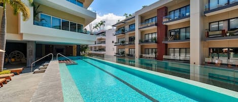 Zona de piscina de hotel con una piscina azul cristalina, sillas de descanso y un edificio moderno con balcones.