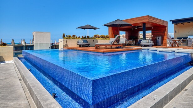 Una piscina en la azotea con sillones de descanso y sombrillas, rodeada por una terraza de madera y vistas al horizonte de la ciudad.