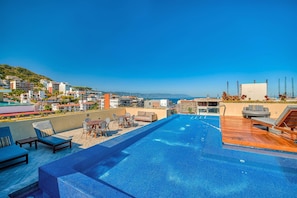 Une piscine à débordement située sur le toit, avec des chaises longues et une vue sur le paysage urbain de la côte.
