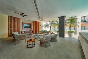 Ein modernes Wohnzimmer mit Flachbildfernseher, bequemen Sitzmöglichkeiten, Deckenventilator und Blick auf Palmen.