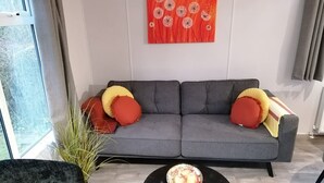 A modern living room with a grey sofa, red and yellow cushions, a black coffee table with fruit, and a large window.