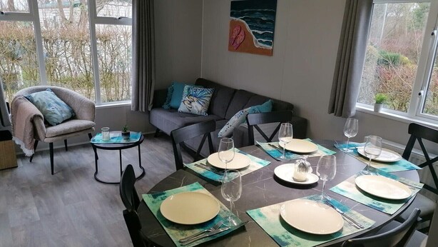 A dining area with a round table set for four, surrounded by chairs. There is a sofa and a chair with a cushion in the background. - Vakantiepark de Zeeuwse Parel - Summerlove (Scherpenisse)