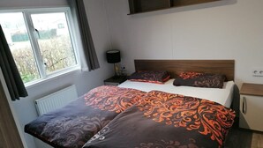 A bedroom with a bed, a nightstand, a lamp, and a window with curtains. - Vakantiepark de Zeeuwse Parel - Summerlove (Scherpenisse)