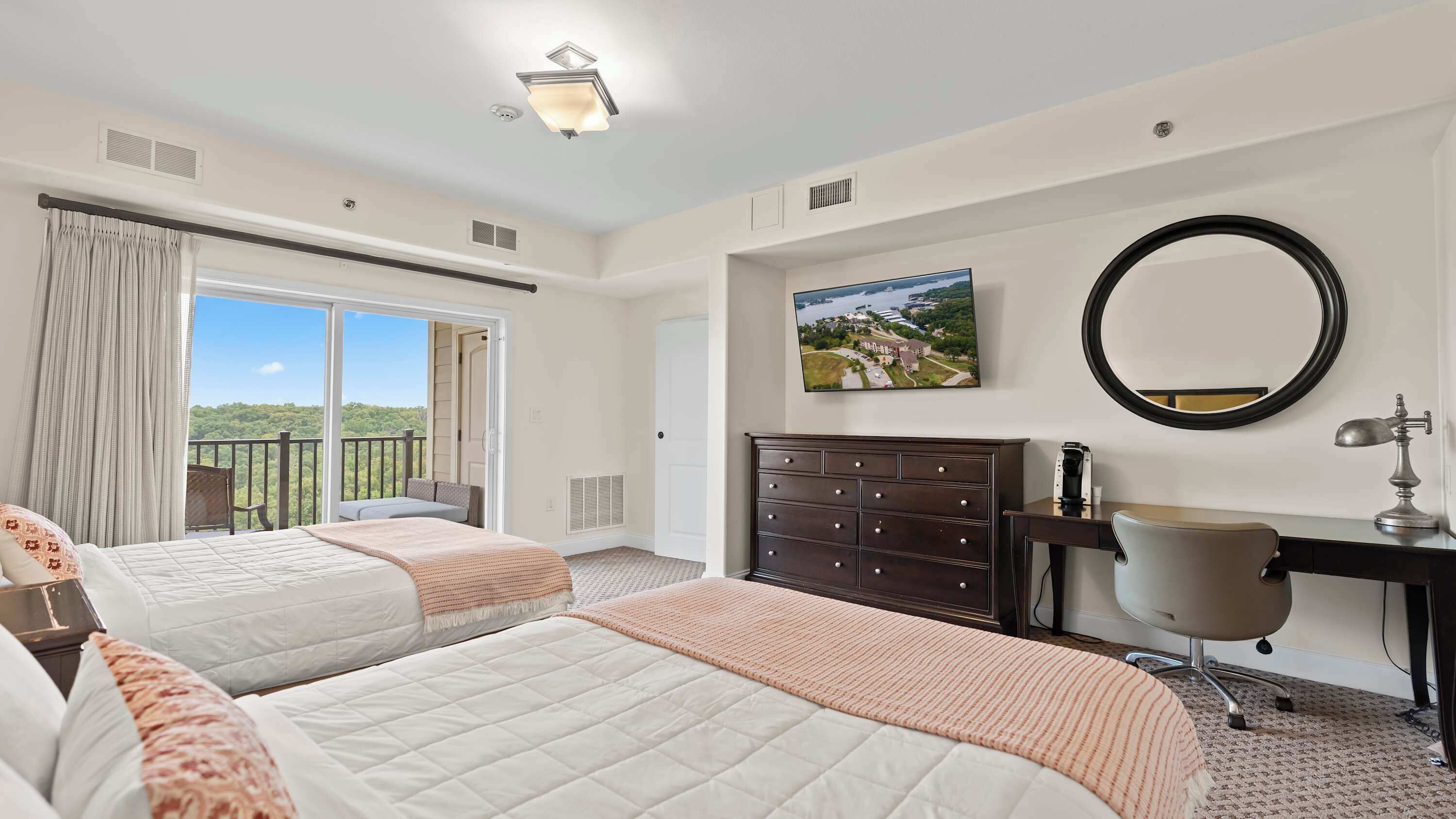 A hotel room with two beds, a desk, a dresser, a mirror, and a window with a view of greenery.
