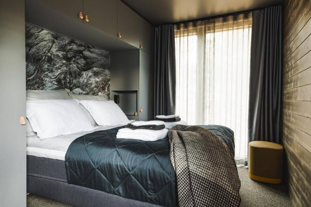 A modern hotel room with a large bed, a headboard with a marble-like design, a bedside table, and a window with curtains.