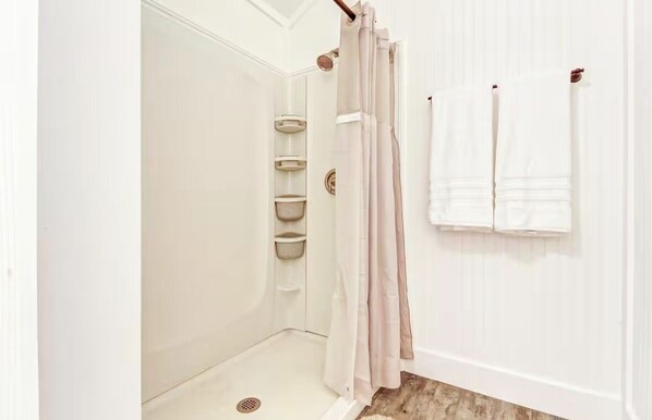 Un baño blanco con ducha, un perchero con dos toallas y un conjunto de estantes con cestas.