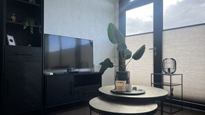 A modern living room with a flat-screen TV, a round table with a vase and candle, a large plant, and a view of the sky through a window.