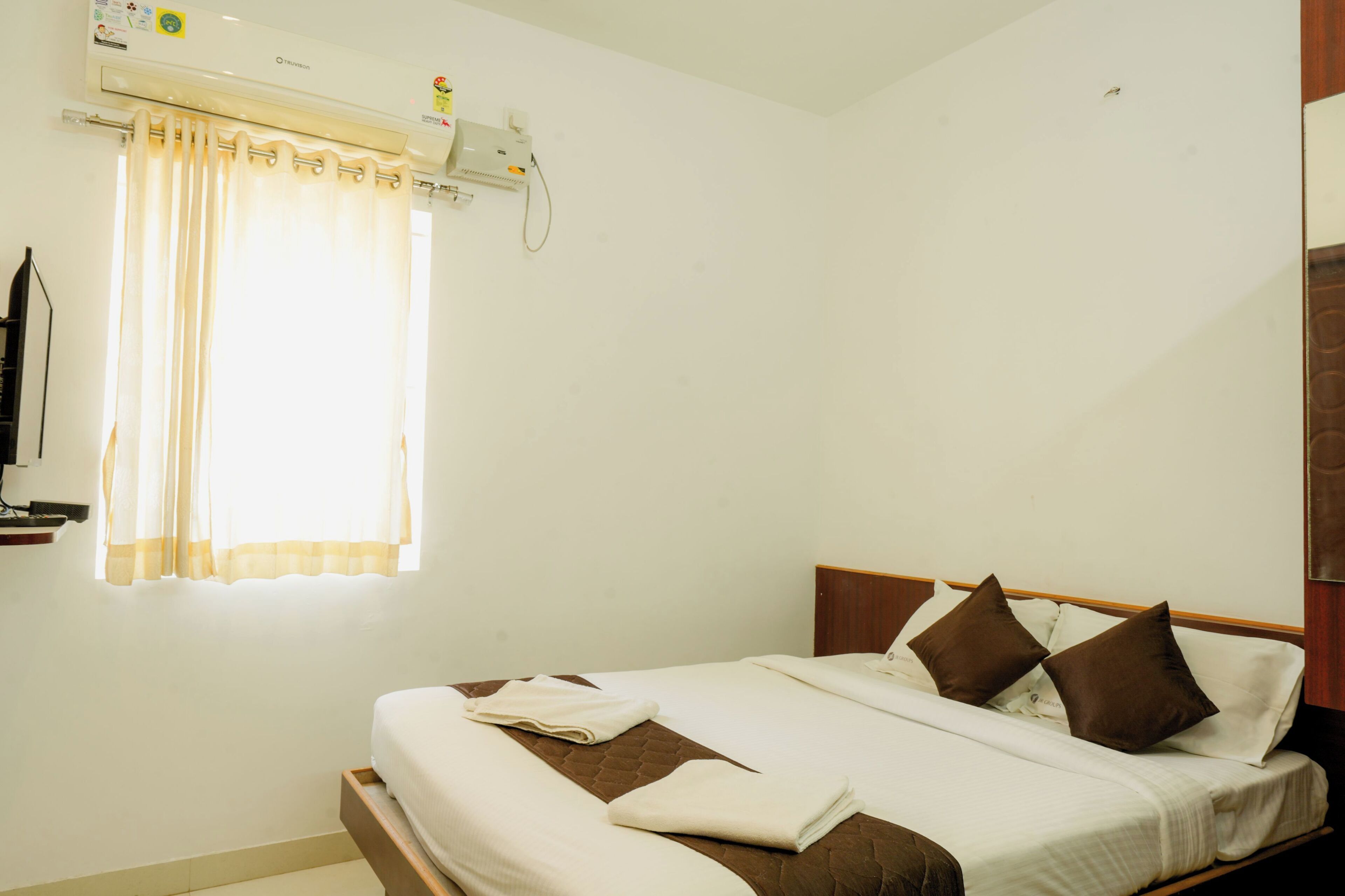 A hotel room with a bed, a window with curtains, an air conditioning unit, and a wall-mounted TV.
