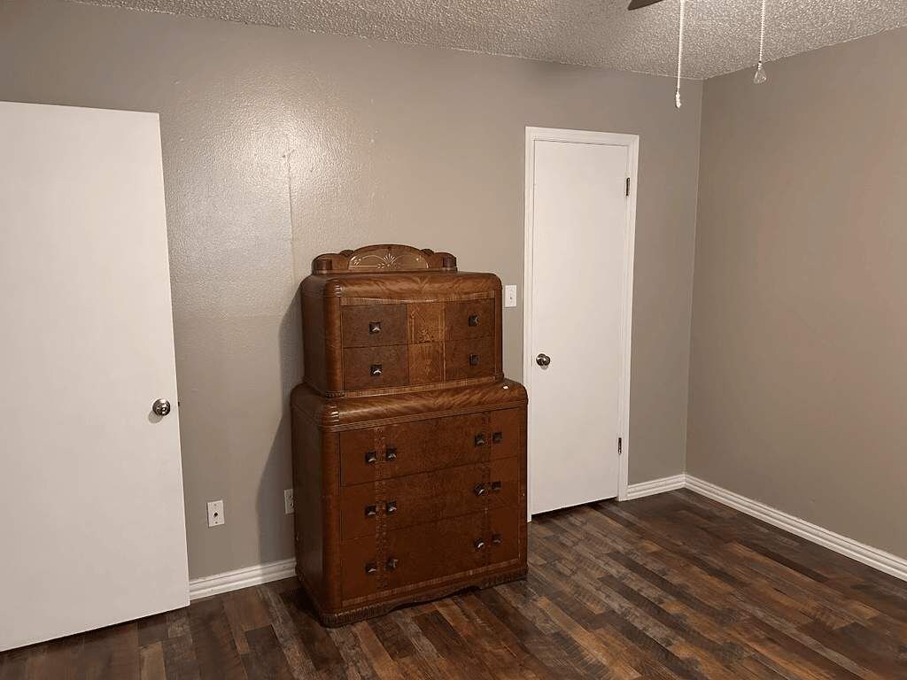 A room with a wooden chest of drawers against a wall, a white door, and a wooden floor.
