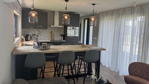 Een moderne keuken met een centraal eiland, barkrukken, hanglampen en een uitzicht op de omgeving door dunne gordijnen.