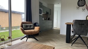 Una sala moderna con una mesa de comedor de madera, un sillón marrón con un cojín negro, y un sillón gris. Se ve una cocina con gabinetes grises y un microondas al fondo.