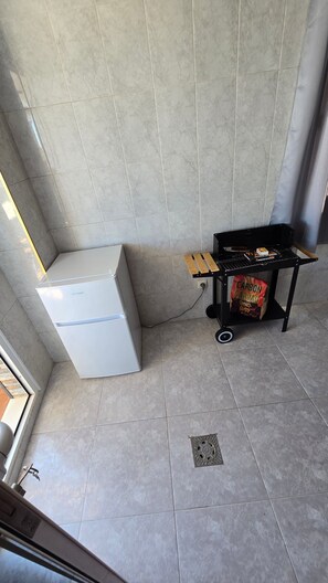 Une petite pièce avec un réfrigérateur blanc, un chariot sur lequel se trouve un sac de charbon de bois et un mur muni d’une grille d’aération.