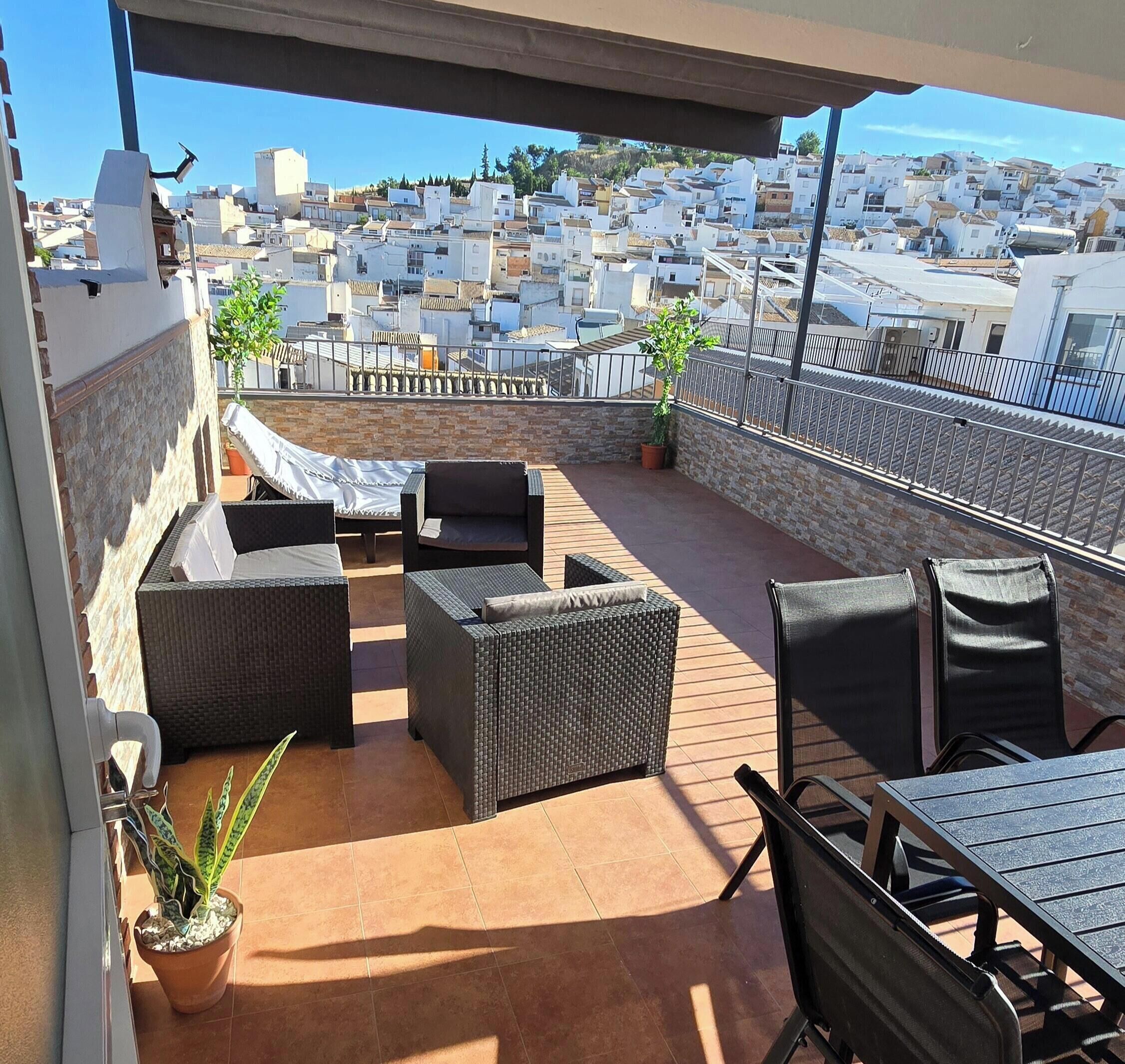 A rooftop patio with wicker furniture, potted plants, and a view of a cityscape.