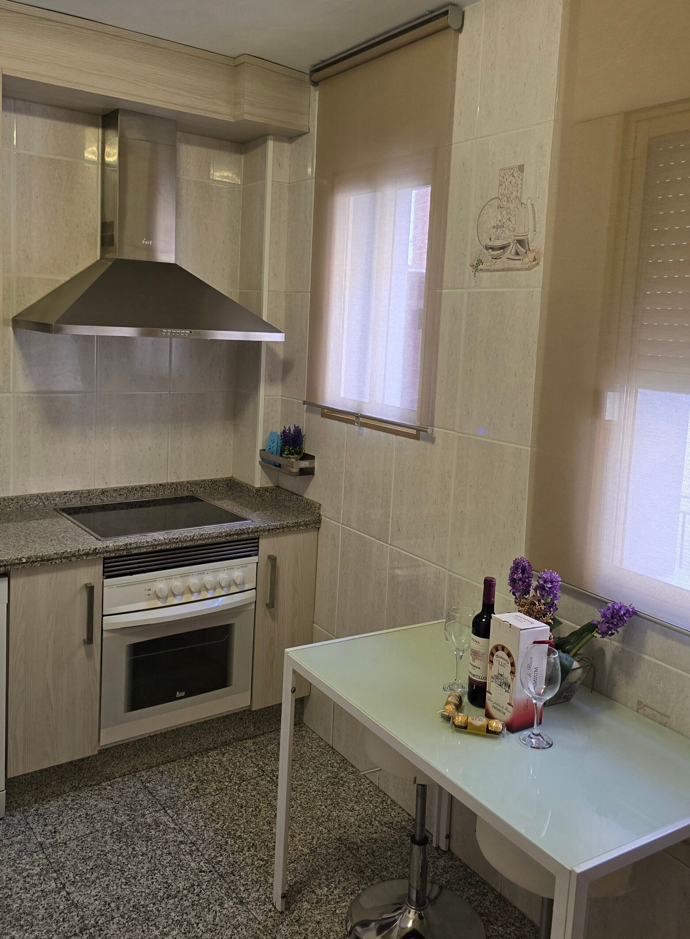 Een keuken met een fornuis, oven en een roestvrijstalen afzuigkap. Er is een raam met zonwering, een klein plankje met decoratieve objecten, en een tafel met een fles en bloemen.