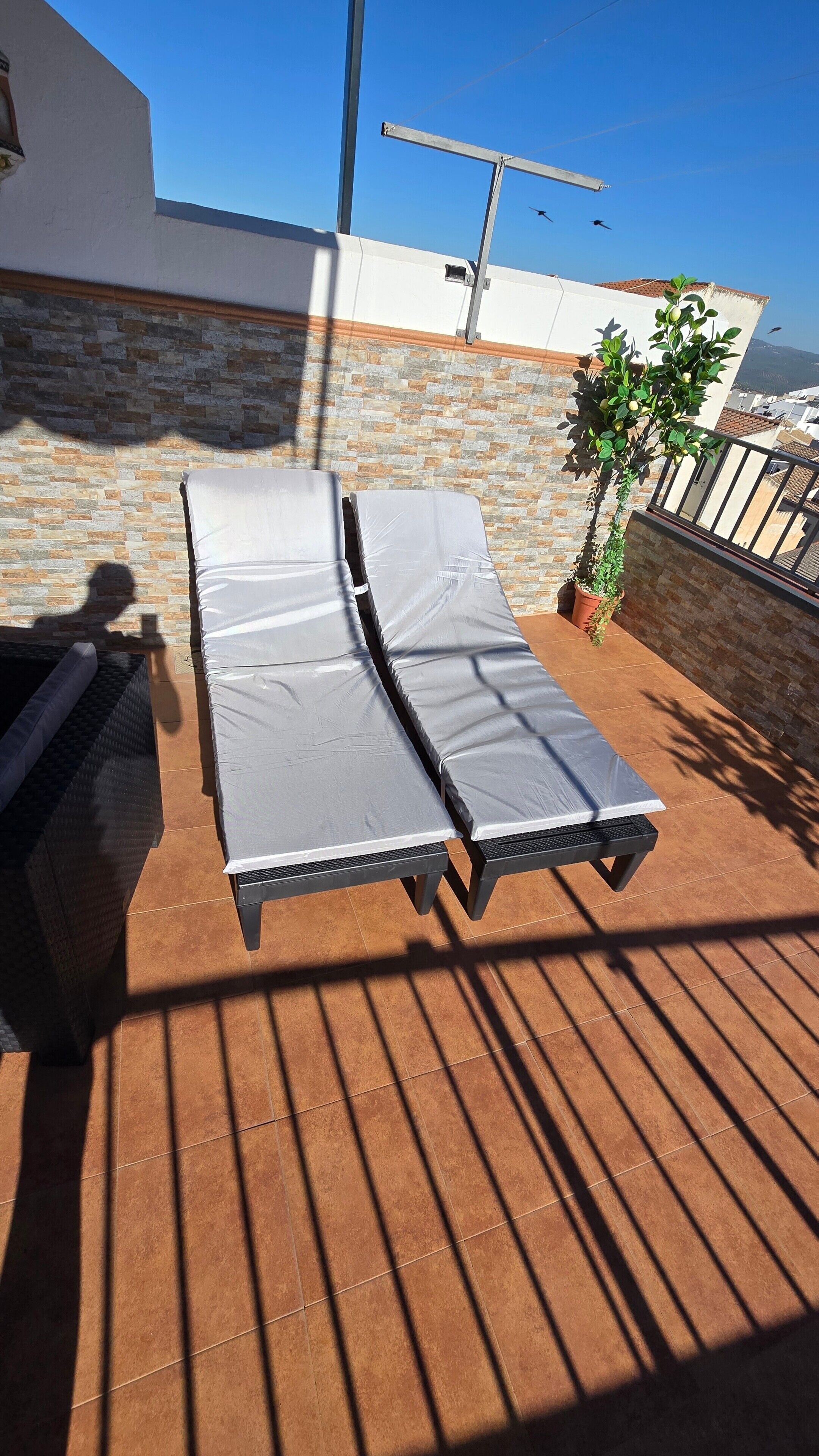 A rooftop terrace with two lounge chairs covered in white, a potted plant, and a view of the cityscape.
