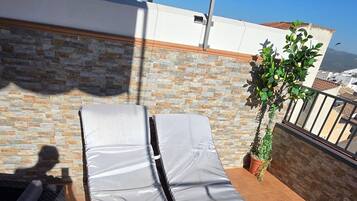 A rooftop terrace with two lounge chairs covered in white, a potted plant, and a view of the cityscape.