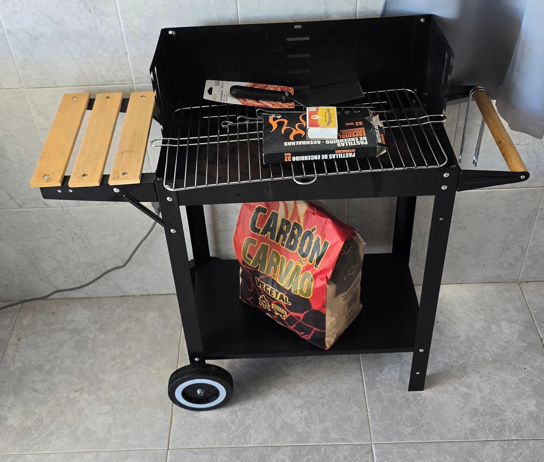 Una parrilla portátil con rejilla metálica, un par de pinzas, una botella de salsa barbacoa y una bolsa de carbón.