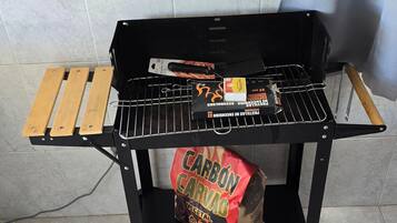 Una parrilla portátil con rejilla metálica, un par de pinzas, una botella de salsa barbacoa y una bolsa de carbón.