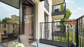Ein Balkon mit Korbstuhl und kleinem Tisch, eine Glastür zu einem Innenbereich und Blick auf die umliegenden Gebäude.