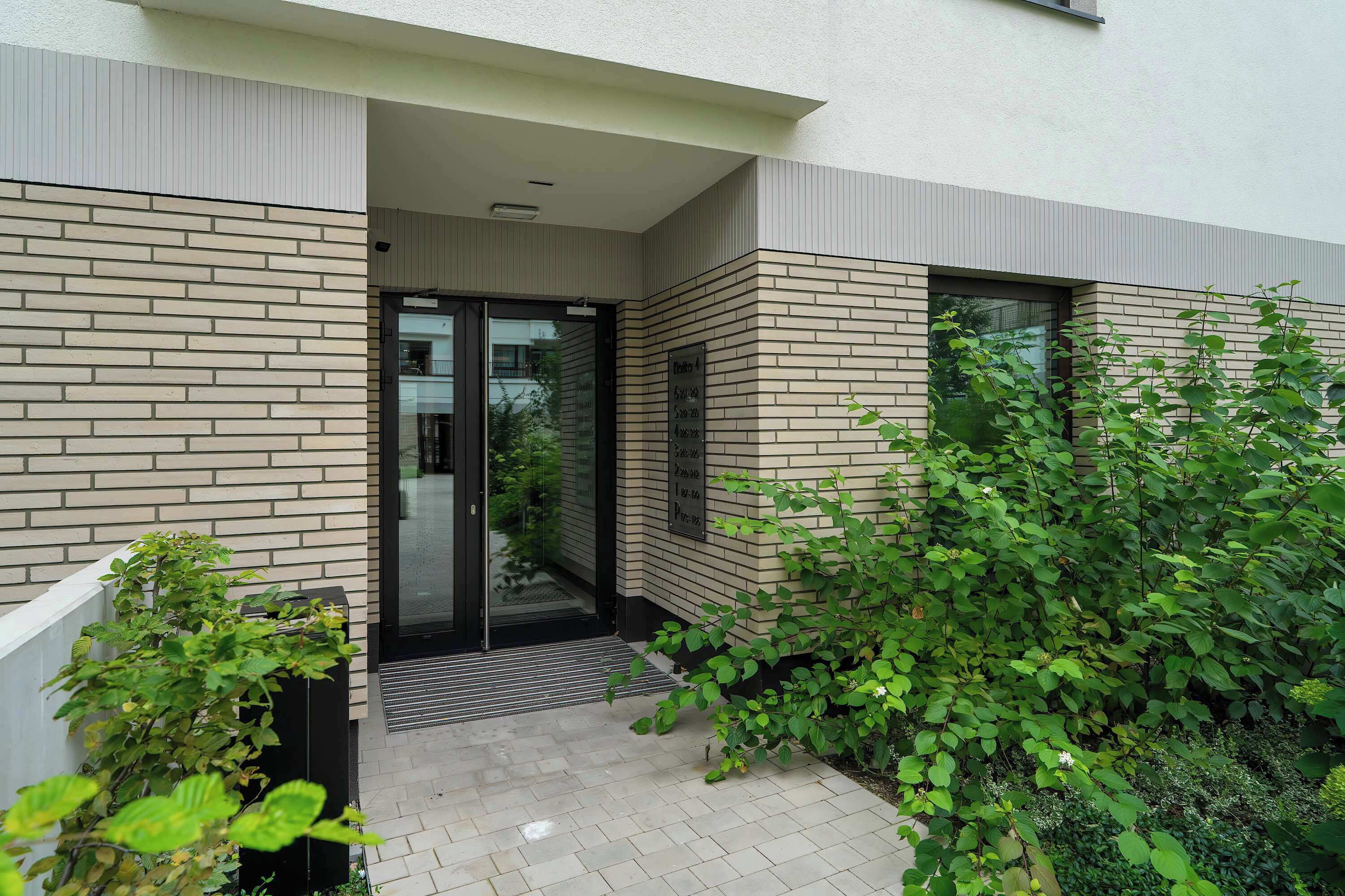 Un bâtiment moderne avec une façade en briques, une entrée en verre et un petit espace jardin agrémenté de plantes vertes.