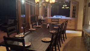 Une salle à manger avec une longue table dressée pour six personnes, entourée de chaises en bois. Un lustre est suspendu au plafond et la pièce est dotée de grandes fenêtres qui offrent une vue sur l’extérieur.