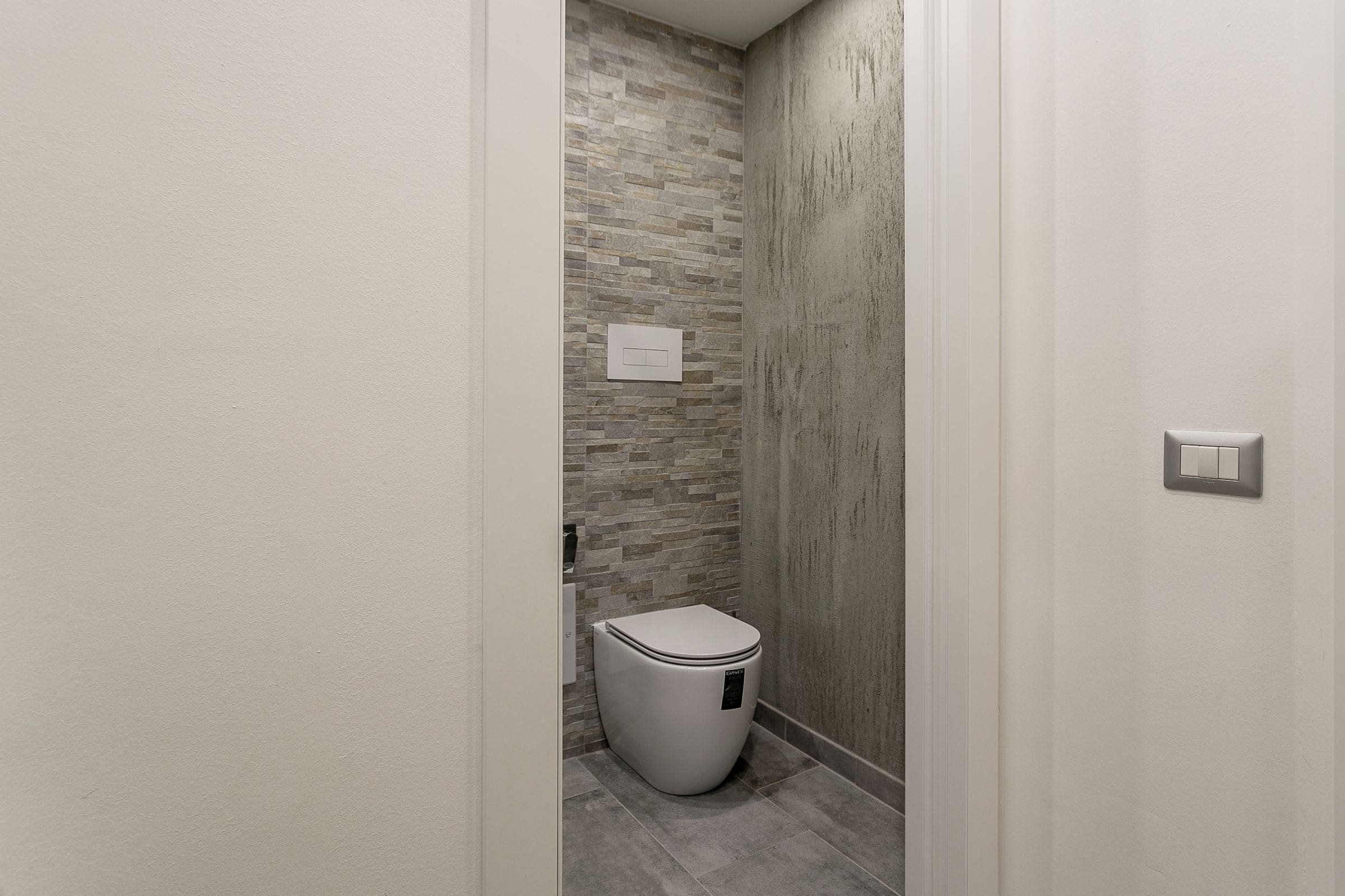 A narrow bathroom with a toilet, a wall-mounted light switch, and a textured stone wall.