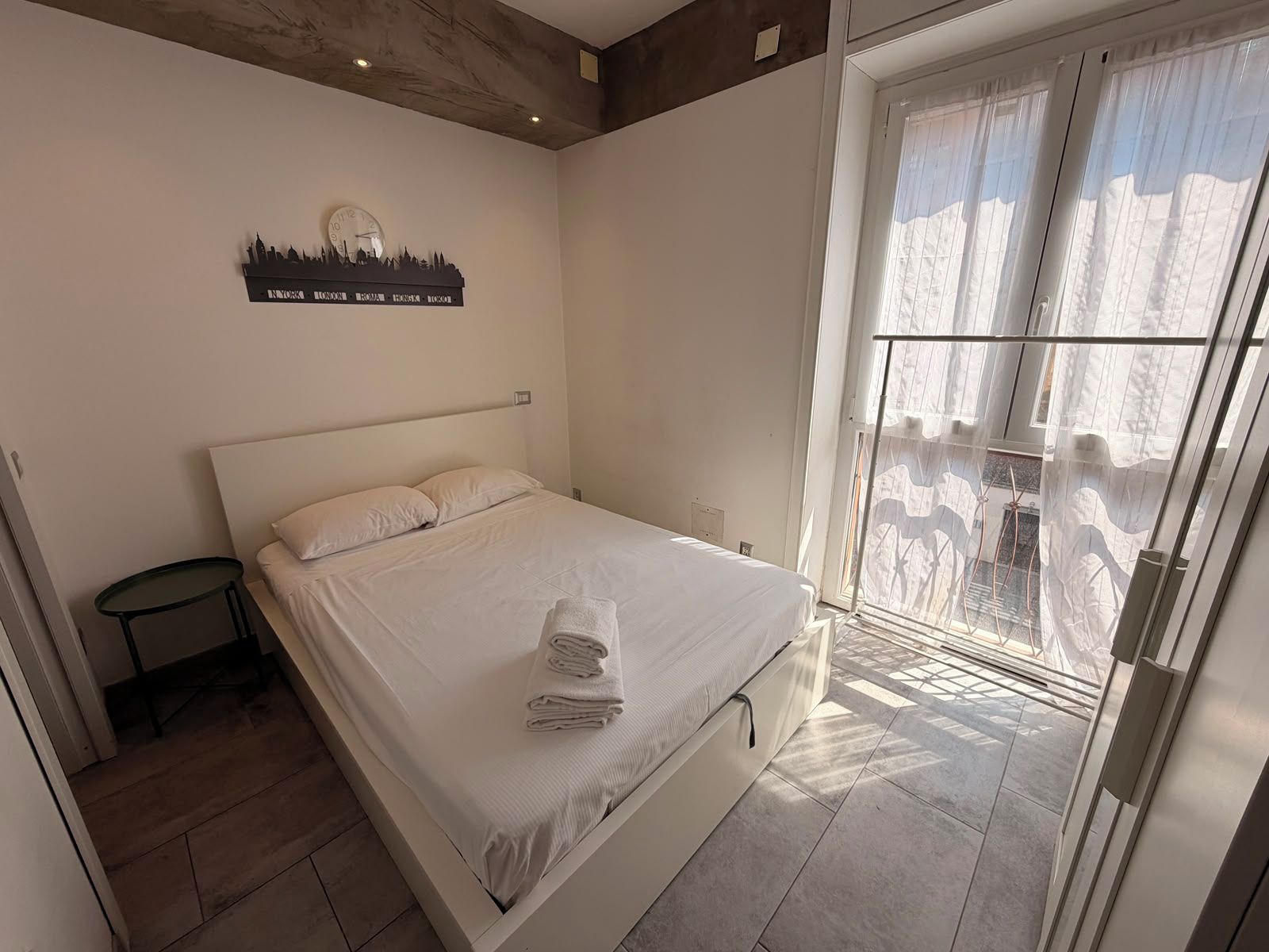 Un dormitorio con cama, mesita de noche, un reloj de pared y una ventana con cortinas.