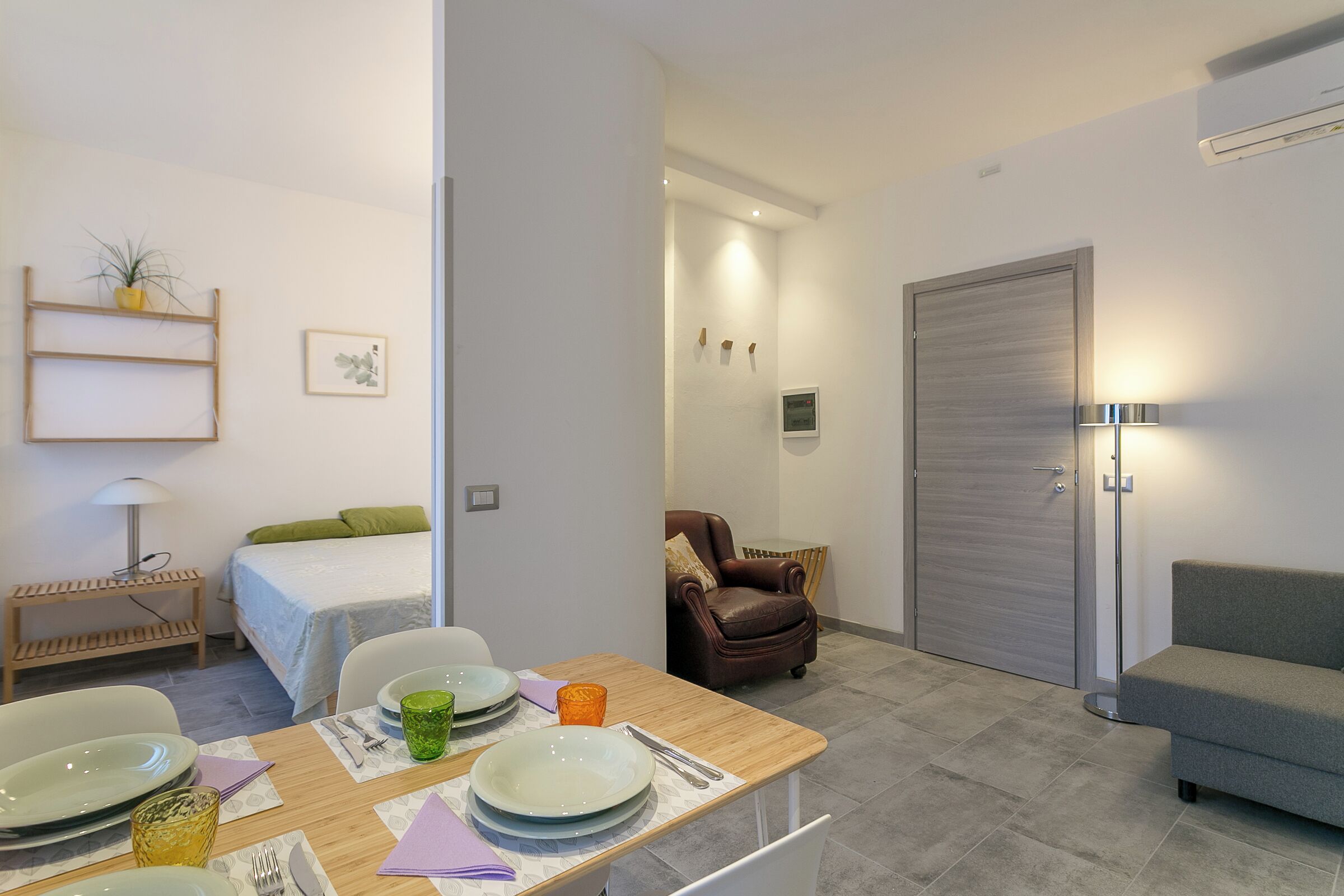 A modern room with a dining table set for four, a bed with green bedding, a brown leather armchair, a grey sofa, and a closed door.
