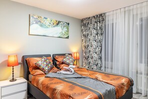 A bedroom with a bed, two bedside lamps, a nightstand, a painting on the wall, and a patterned curtain.
