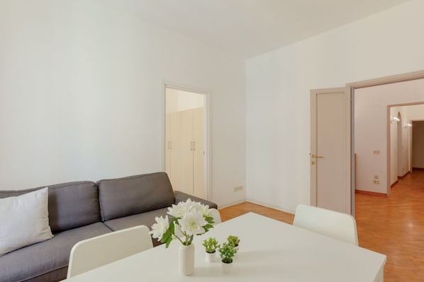 Un salon moderne avec un canapé gris, une table blanche ornée d’un vase de fleurs et une porte donnant sur une autre pièce.