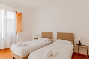 Une chambre avec deux lits, une porte en bois, une petite table avec une lampe et une fenêtre avec des rideaux légers.