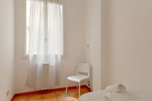 Une pièce avec une chaise blanche, un lit sur lequel est posée une serviette blanche enroulée, une fenêtre avec des rideaux légers et un sol carrelé.