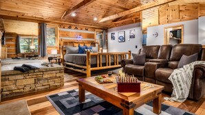 Un interior de cabaña acogedor con chimenea de piedra, vigas de madera en el techo, un sofá, un sillón de cuero y un tablero de ajedrez sobre una mesa de centro.