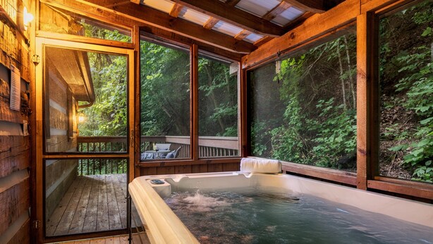 Una cabaña de madera con jacuzzi, amplias ventanas y una terraza con vistas a un bosque.
