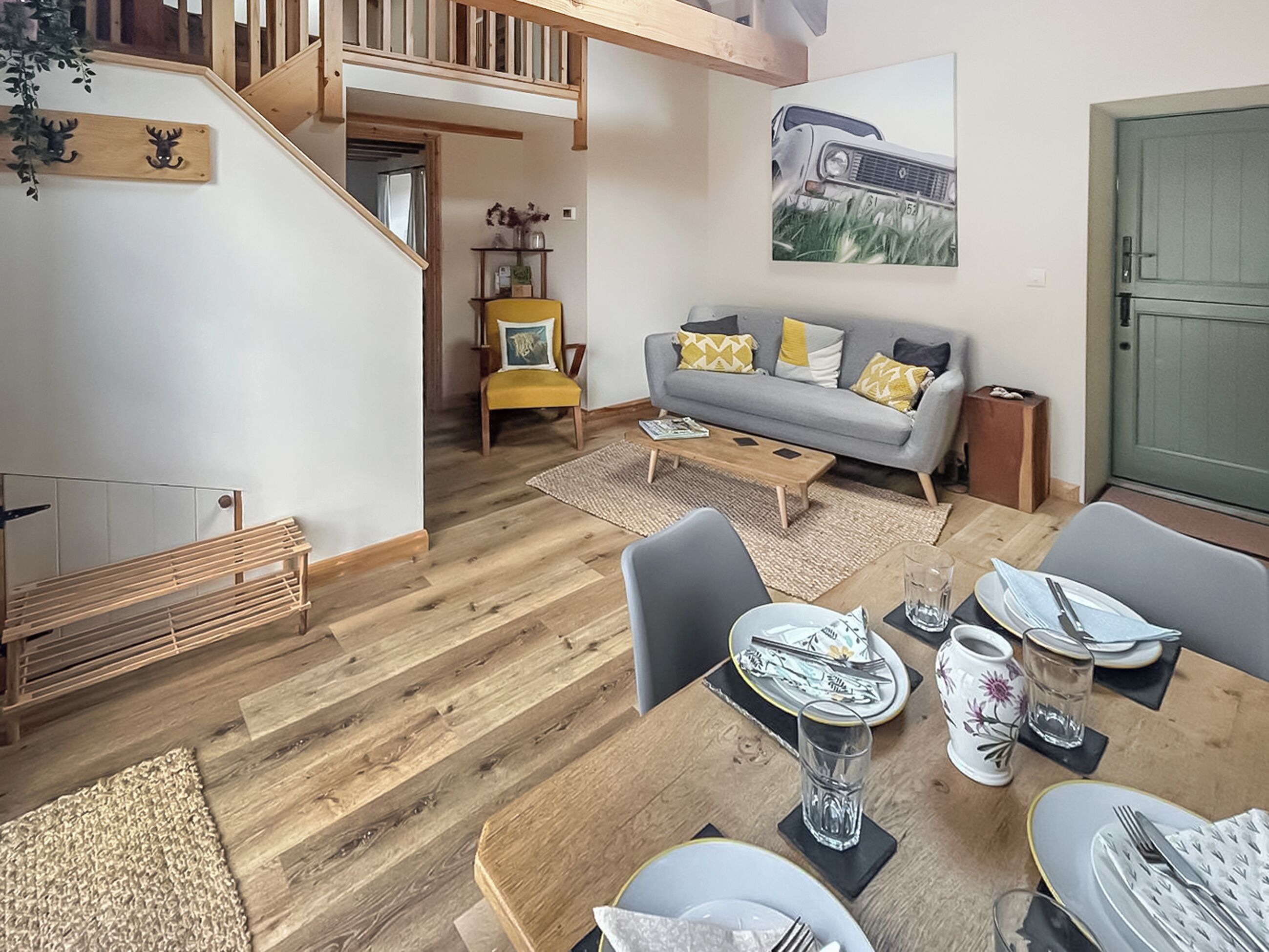 A spacious living area with a dining table set for four, a sofa with cushions, a staircase, and a painting on the wall.