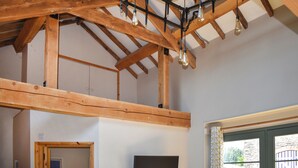 A room with exposed wooden beams, a ceiling light fixture, and a window with a view of greenery outside. - The Calving Barn - UK49679 (Bratton Fleming)