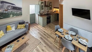 A modern living room with a grey sofa, a dining table set for four, and a kitchen in the background. - The Calving Barn - UK49679 (Bratton Fleming)