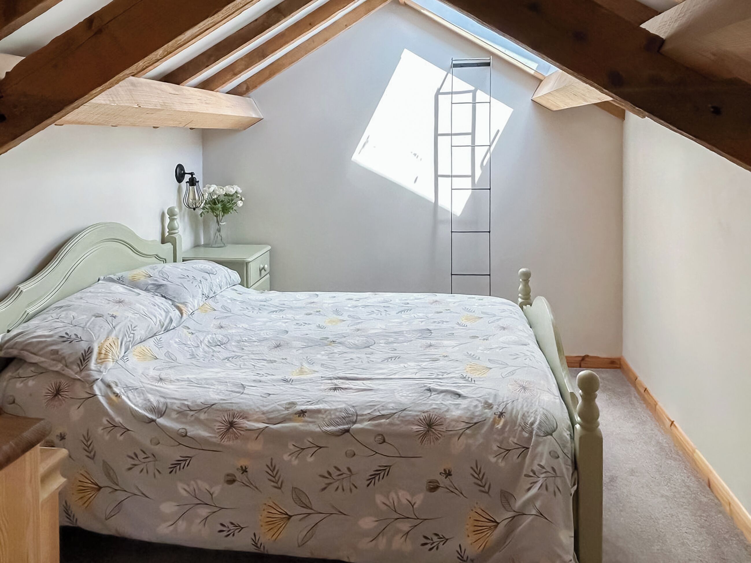 A bedroom with a sloped ceiling, a bed with a floral bedspread, a nightstand with a vase of flowers, and a skylight.