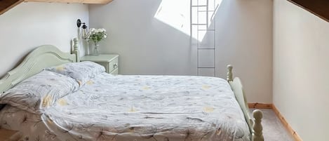 Un dormitorio con techo inclinado, una cama con edredón floral, una mesita de noche con un jarrón de flores y un tragaluz.