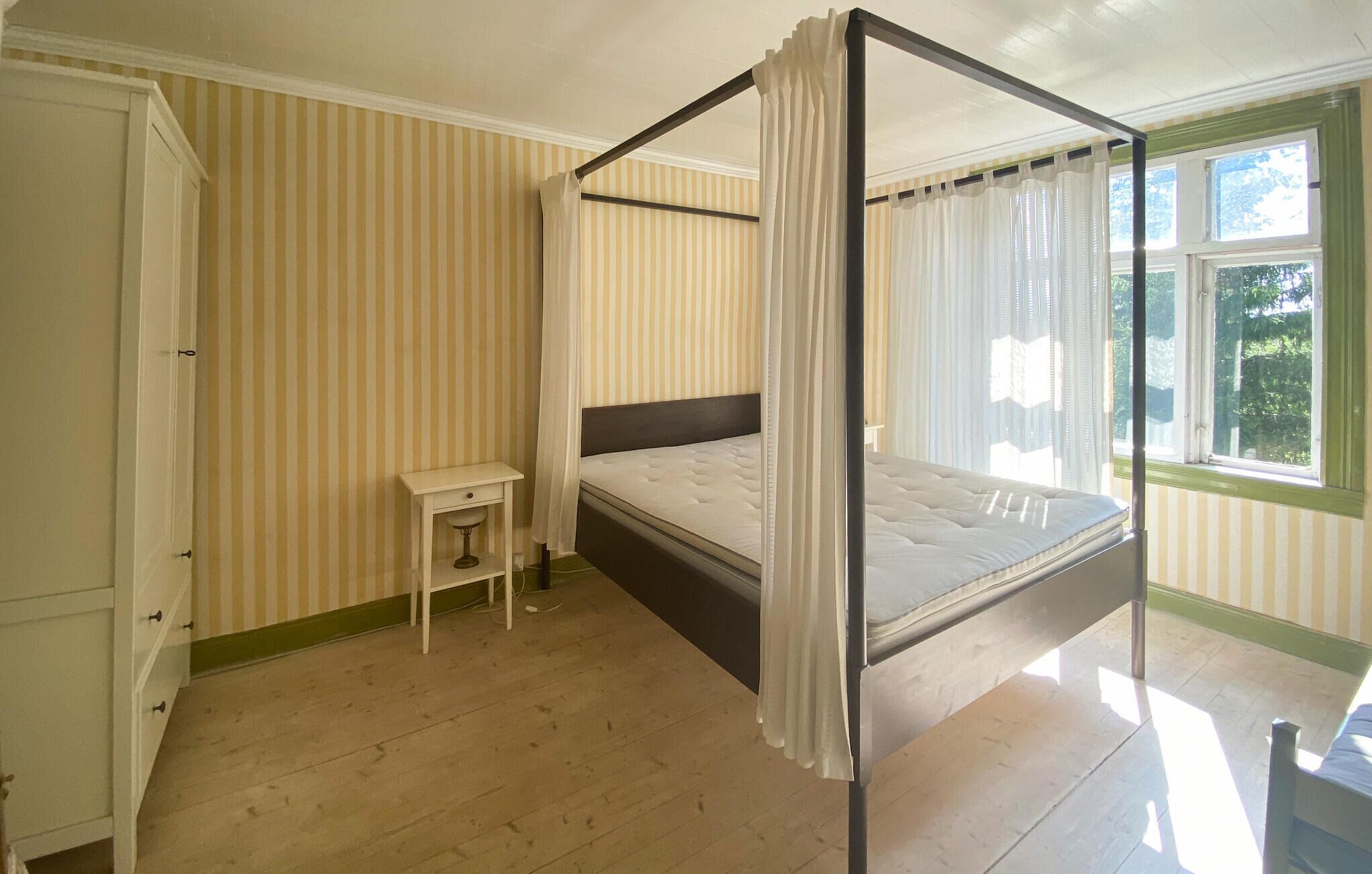 Una habitación con una cama con dosel, una mesita de noche pequeña, un armario y una ventana con cortinas.