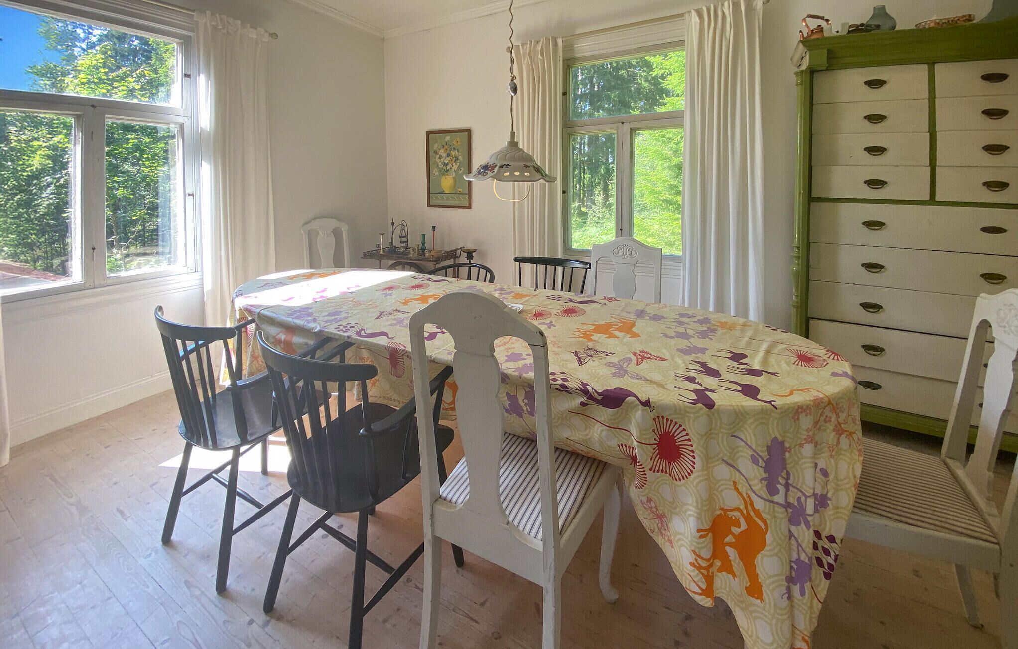 Ein Esszimmer mit einem für vier Personen gedecktem Tisch, umgeben von Stühlen, einer Kristallleuchtre und einer grünen Kommode.