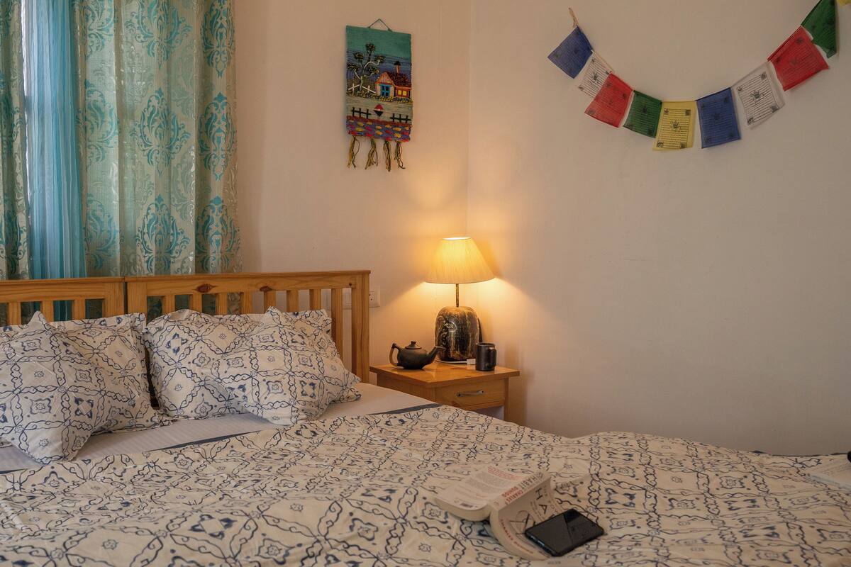 A neatly made bed with patterned bedding, a bedside table with a lamp, a wall hanging, and a window with curtains.