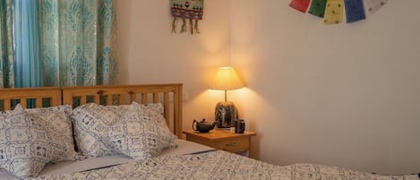 Ein ordentlich bezogenes Bett mit gemusterten Betttextilien, ein Nachttisch mit Lampe, eine Wanddekoration und ein Fenster mit Vorhängen.