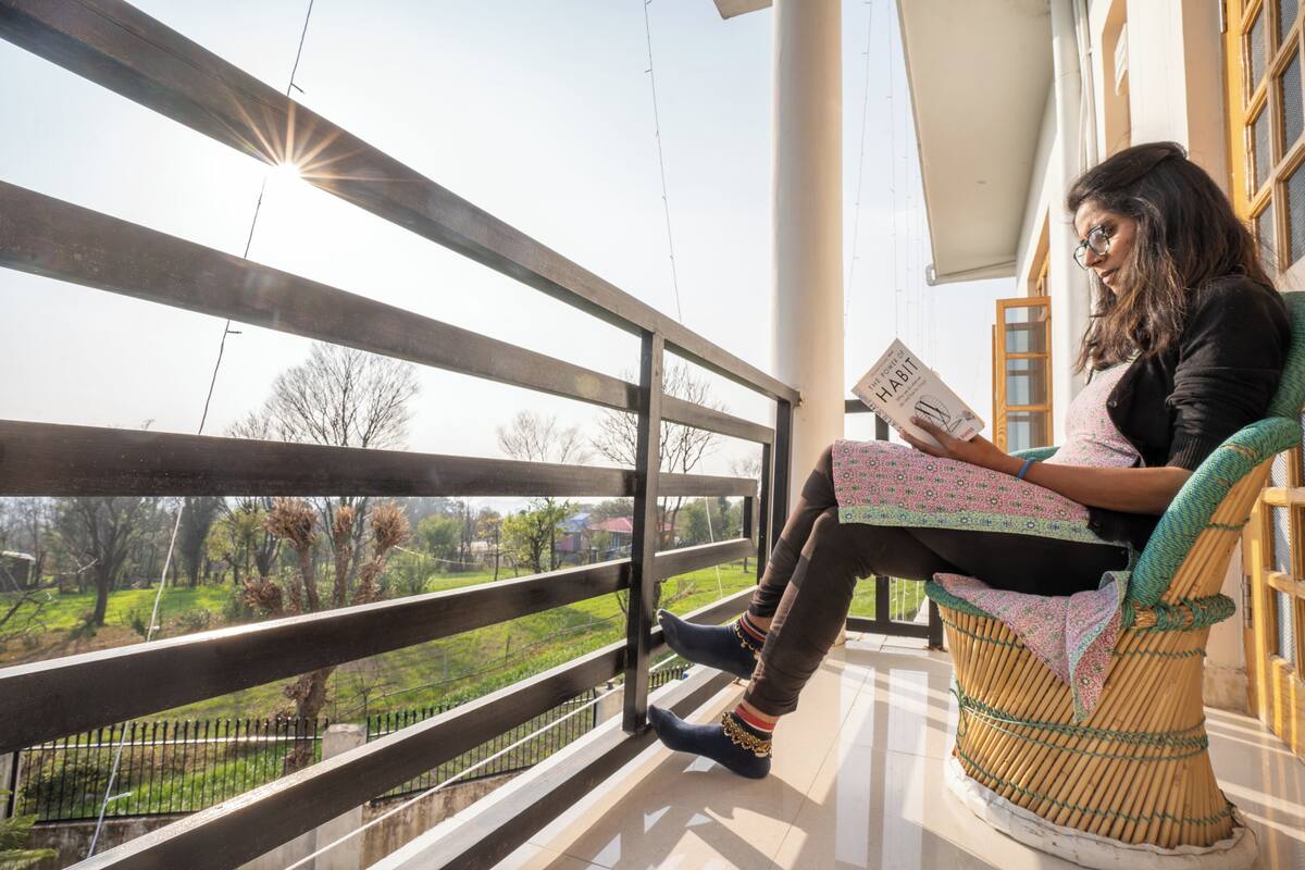 A person sitting on a wicker chair on a balcony with a view of a park and buildings in the background.