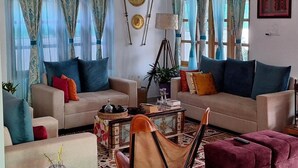 Ein Wohnzimmer mit zwei Sofas, einem Couchtisch und einem roten Hocker. Vorhänge, eine Lampe und dekorative Teller sind an der Wand angebracht.