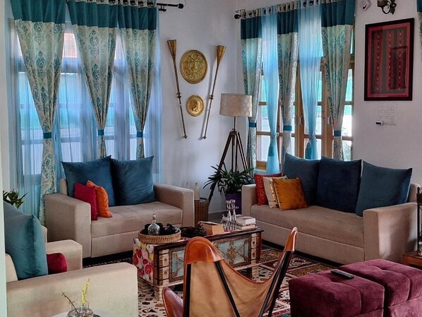 A living room with two sofas, a coffee table, and a red ottoman. There are curtains, a lamp, and decorative plates on the wall.